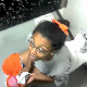 A black girl is video-recorded by her friends sitting on a toilet in a public restroom. She may be peeing or pooping. There are no sounds to indicate which.